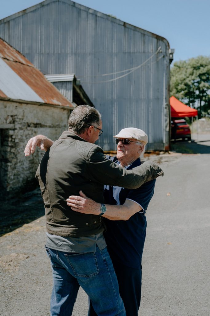 Two men embrace outside near a grey barn on a sunny day.