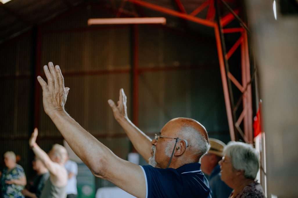 Elderly people raising their hands inside a rustic barn setting.