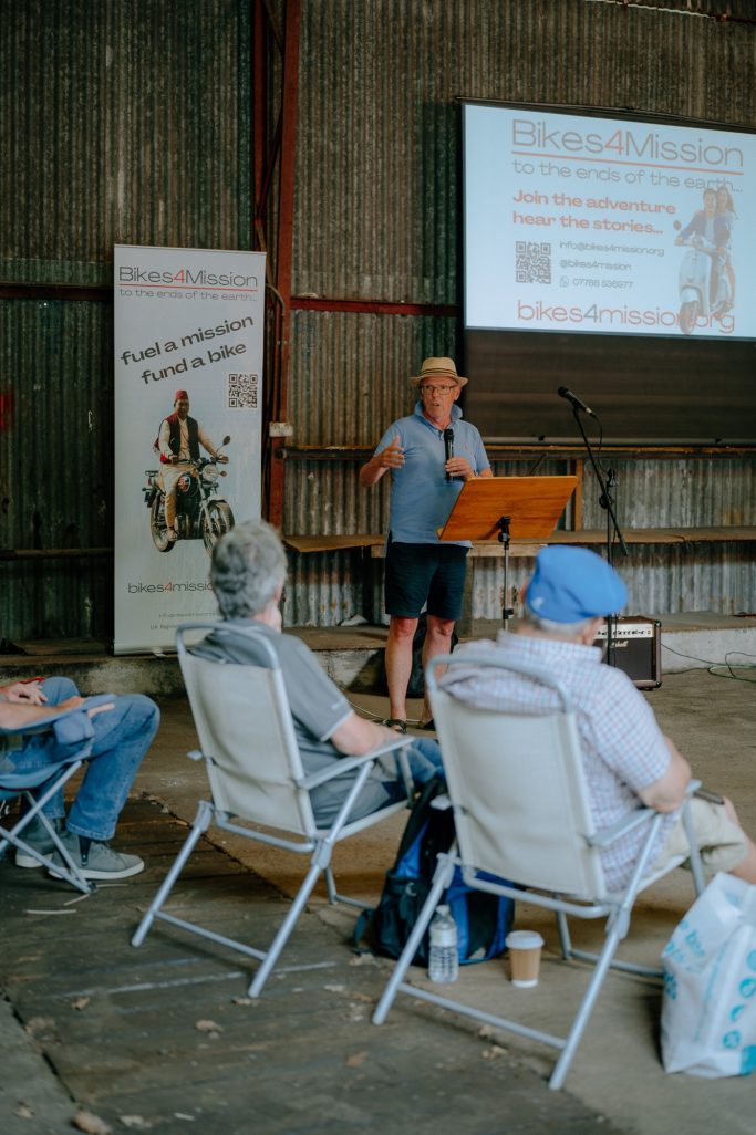 A speaker addresses an audience seated in chairs with a presentation screen behind.