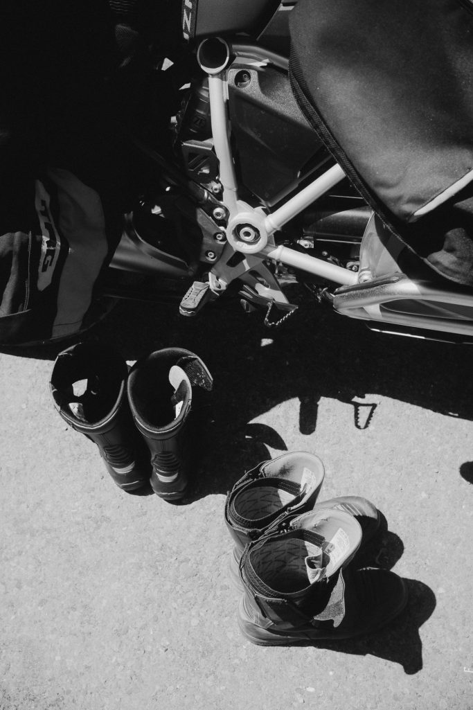 Pair of black boots next to a motorcycle on a paved surface.