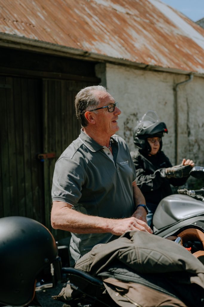 Man wearing glasses, seated with a motorcycle in the background and a woman preparing nearby.