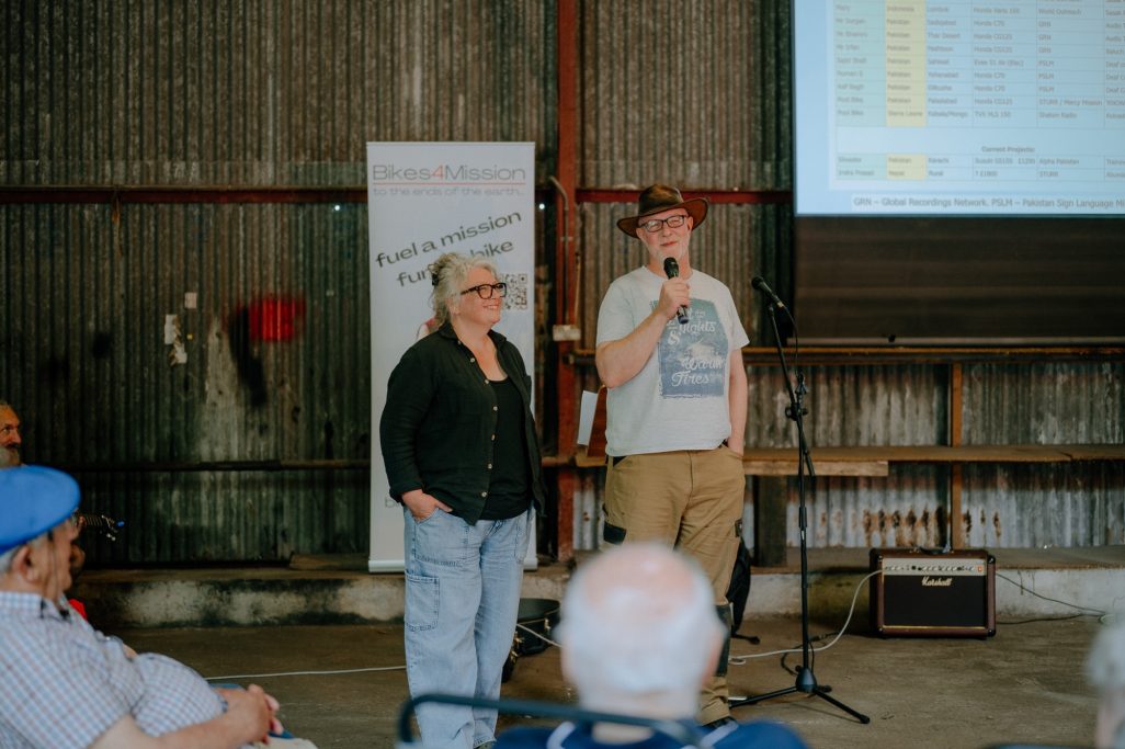 Two people speaking at a microphone in an industrial setting with an audience.