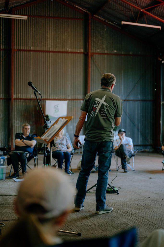 A presenter speaks to a seated audience in a rustic venue.
