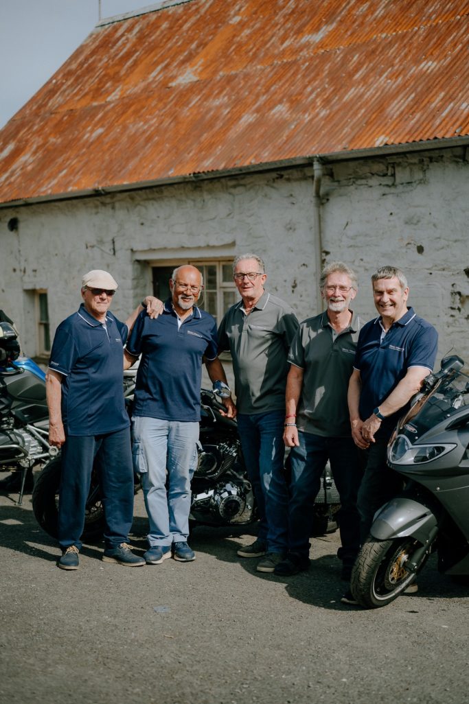 A group of five men standing beside motorbikes in front of a rustic building.