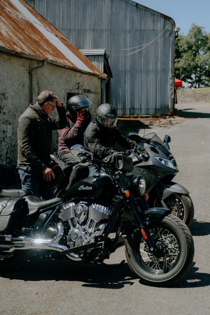Two motorcyclists in helmets beside their bikes, with a man standing nearby.