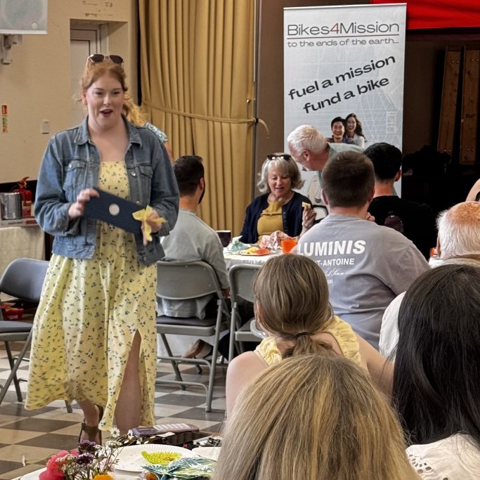 A woman in a yellow dress speaks to an audience at a social event with refreshments.