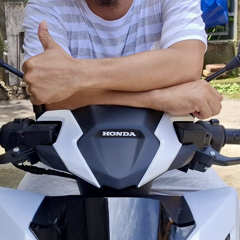 A person smiles while sitting on a red scooter, surrounded by several smiling villagers.